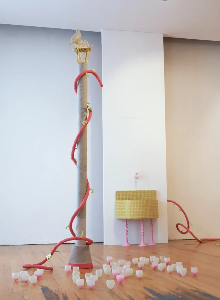 Photographie d'une installation d'art contemporain dans une galerie au parquet de bois. Un grand candélabre gris texturé est entouré d'une vigne rouge et verte sculptée, surmonté d'une lanterne dorée et d'un pigeon blanc. À droite, un meuble-vasque jaune à damier jaune avec un robinet rose est posé contre un mur blanc. Au sol, de nombreuses coupes ou verres blancs avec une bordure rose sont dispersés.