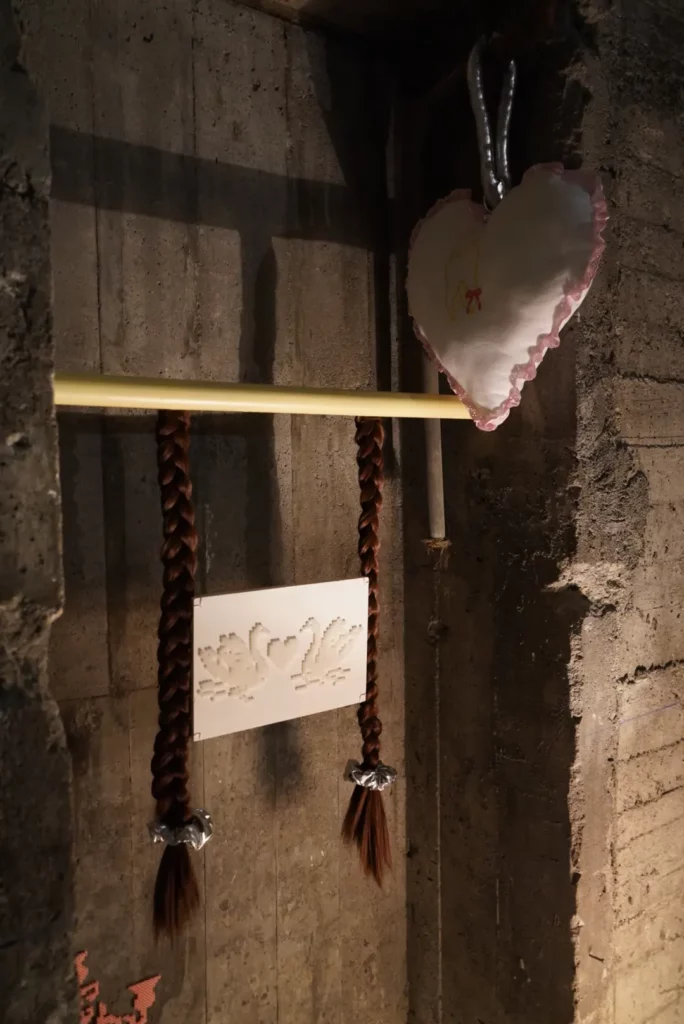 Installation artistique contemporaine montrant deux longues tresses de cheveux bruns suspendues à une barre jaune horizontale. Elles encadrent un panneau rectangulaire en dentelle blanche orné de deux cygnes et d'un cœur entrelacés. À droite, un grand cœur en tissu froncé blanc et rose est suspendu par un crochet métallique contre un mur en béton brut.