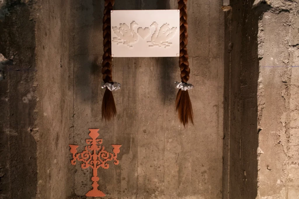 Installation artistique contemporaine sur mur de béton brut : deux longues tresses de cheveux synthétiques bruns, terminées par des élastiques argentés froissés, suspendent un panneau de dentelle blanche représentant deux cygnes et un cœur entrelacés. En bas à gauche, un motif d'ornementation baroque ou chandeliers en perles Hama orange est posé au sol.