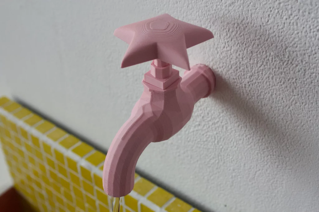 Photographie rapprochée d'un robinet d'art contemporain de couleur rose pastel mat, fixé sur un mur blanc texturé. Sa poignée est remplacée par une étoile à cinq branches sculptée. Le corps du robinet a des formes géométriques facettées. En bas à gauche, une portion de mur ou d'étagère est revêtue de petites mosaïques carrées jaunes.